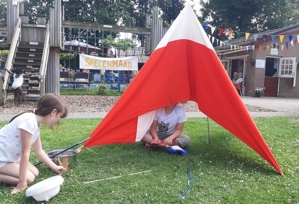 Punttentje in rood en wit met daarin een kind. Naast de tent zit een meisje. Achter het tentje zie je het houten klimfort met het spandoek van SpelenMaaR.