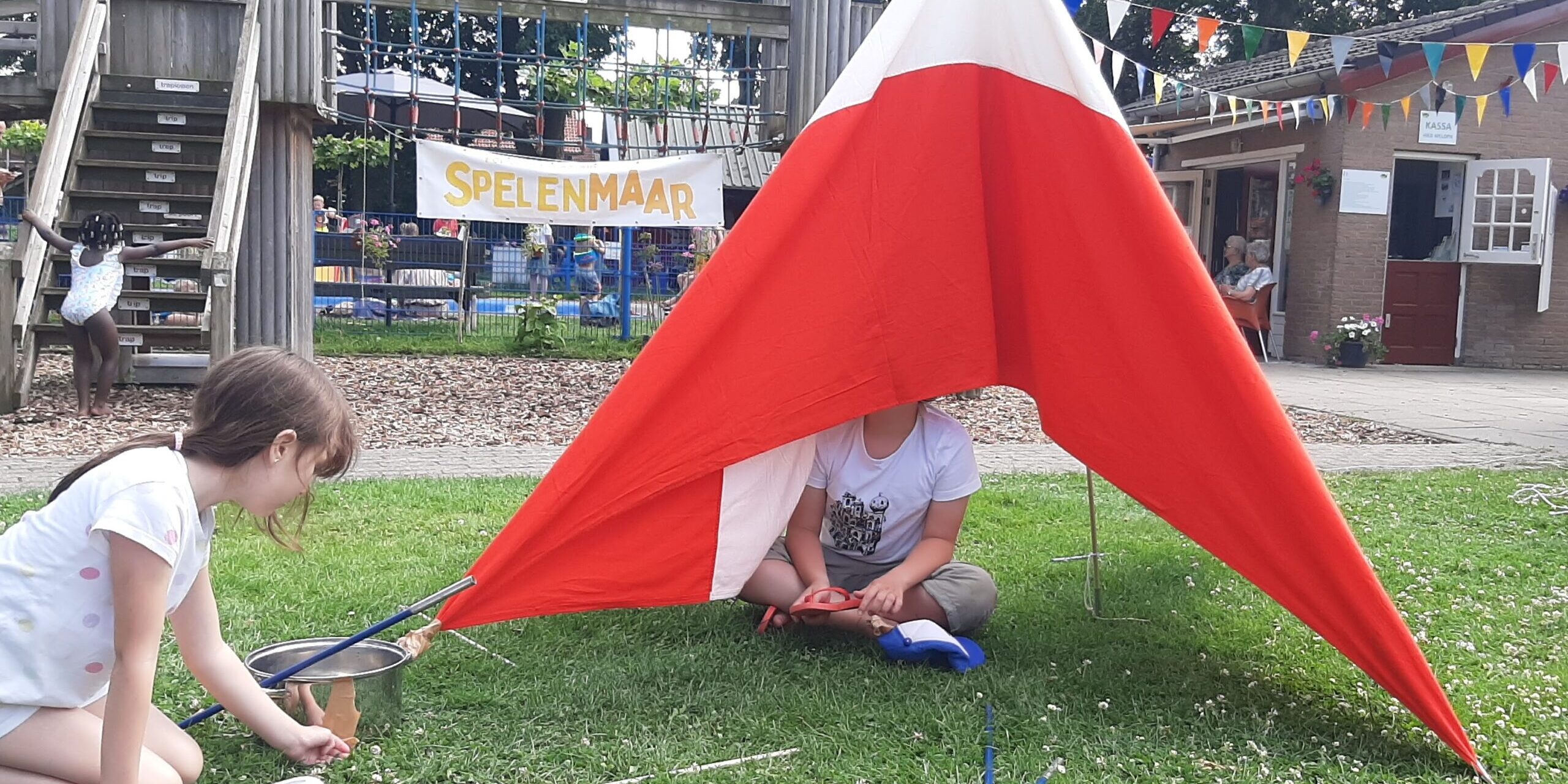 Punttentje in rood en wit met daarin een kind. Naast de tent zit een meisje. Achter het tentje zie je het houten klimfort met het spandoek van SpelenMaaR.